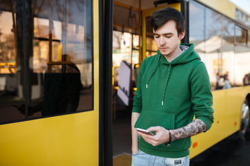 Portrait of young man with dark hair standing outside with mobile phone in hand and bus on background. Thoughtful boy in green sweatshirt standing on street and looking in his cellphone