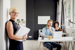 © chika_milan - Senior standing in modern office against the wall and holding notebook