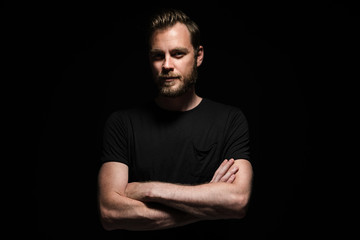 portrait of a bearded man with an intense look standing in a dark room against a black background.