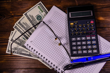 Notepad, pen, calculator and dollar bills on wooden table. Top view