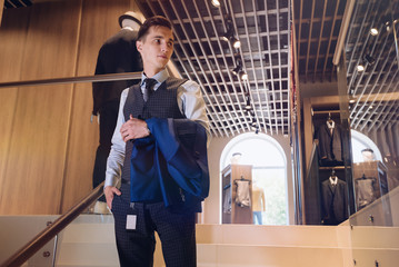 Wall Mural - elegant guy in a business suit in a clothing store is holding a suit with a price tag standing on the stairs