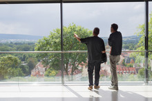 Two Business Man Looking Out Of Modern Office Window Pointing With Arm, From Back