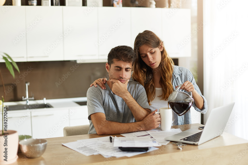 Picture of frustrated young European family working through finances in kitchen, calculating expenses, trying to save some money; caring wife serving coffee to her stressed husband deep in thoughts