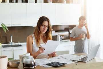 Wall Mural - Busy female worker working with pile of documents, reading papers with attentive look, studying diagrams and charts, being absorbed into work while her husband chatting over mobile phone, drinking tea