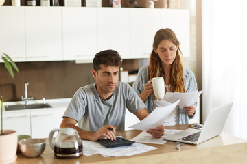 Wall Mural - Picture of serious young American couple spending weekend morning in kitchen, doing paperwork, looking through mail and calculating family expenses, paying bills online using laptop computer