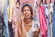 Upset woman standing near rack of clothes, chatting over smart phone with her friend, complaining that she has nothing to wear. Displeased female not knowing what to put on for birthday party