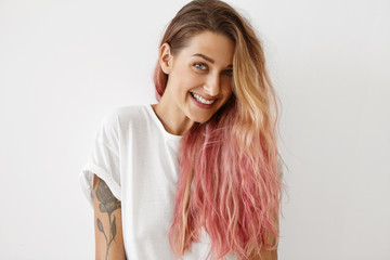 Wall Mural - Style, fashion and hair coloring concept. Portrait of gorgeous young female with charming smile, tattoo on arm and long loose hair dyed pink looking at camera, having joyful carefree expression