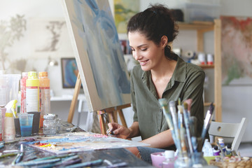 Picture of cute female artist sitting at table, surrounded with watercolors, drawing something at easel, having happy expression. Brunette young woman being busy with creative work at workshop