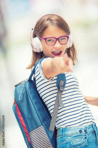 Zdjęcie XXL Uczennica z torbą, plecakiem. Portret nowożytna szczęśliwa nastoletnia szkolna dziewczyna z torba plecaka hełmofonami i pastylką. Dziewczyna z aparatami ortodontycznymi i okulary.