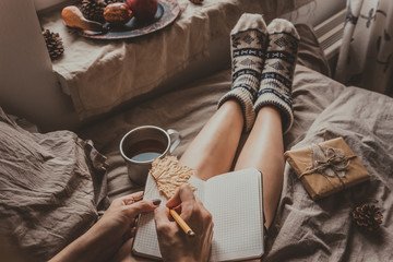 Cozy home. Woman with cup of hot drink sitting by the window and writing in notepad