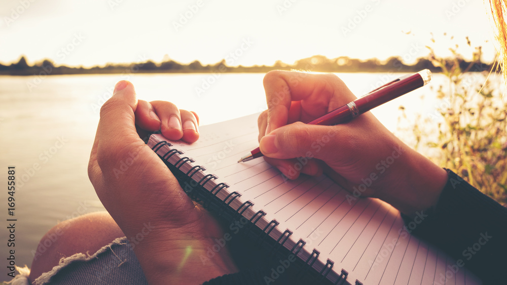 Close up hand of young woman with pen writing on notebook at riverside in the evening. - obrazy, fototapety, plakaty 