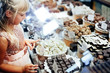 A little blonde girl is standing near a shop window with an assortment of sweets. The child chooses sweets