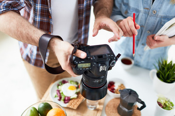 Close-up shot of hard-working food photographers looking through made photos on camera, served table for breakfast with fresh fruits, herbal tea, fried egg and lettuce