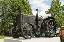 An Old Black Wheeled Tractor Stands As A Monument On A White Pedestal Closeup