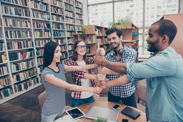 Team work concept. Four happy international young students putting their fists on top of each others, finished preparing their project, standing in the book store, wearing casual clothes