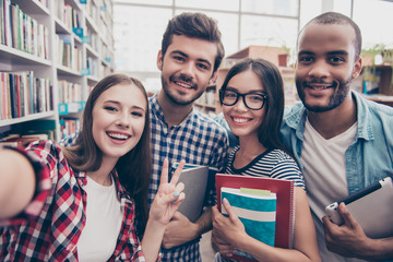 Wall Mural - Selfie time! Four international students with beaming smiles are posing for selfie shot, caucasian attractive lady is taking, in school library building. Gathered, cheerful, smart and successful youth