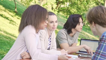 Canvas Print - Multiethnic group of young students friends sitting and talking outdoors while using laptop computer. Looking aside.