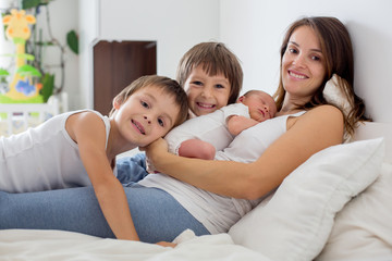young mother lying in bed with her newborn baby boy and his older brothers