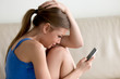 Stressed young woman crying sitting on sofa at home reading message with bad news holding cell phone. Upset lady frustrated after breakup, terrible loss, problems in hopeless relations with boyfriend