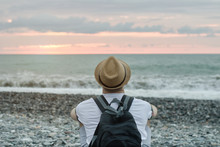 Young Man In Hat And With Backpack Sitting On The Beach Against The Backdrop Of The Sea And Sunset Sky. Back View