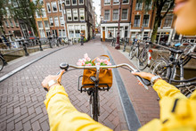 Woman Riding A Bicycle With Bouquet Of Flowers On The Street In Amsterdam City. View On The Hands Holding Helm