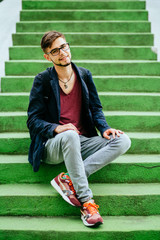 Portrait of a handsome teen student guy in glasses sitting on the green sairs - education, hischool and people concept.