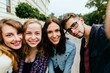© Iryna - Focus on four positive student friends taking selfies and posing arm in arm during univercity park background .They are wearing summer clothes, guy in glasses.