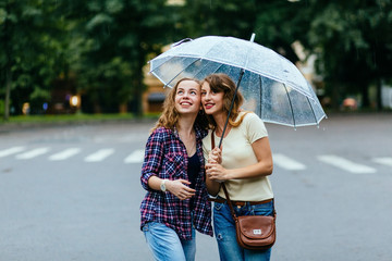 Two joyful hipster girl sheltering from rain beneath umbrella - they loves a rain and rejoices. Large rain drops fall on an umbrella dome.