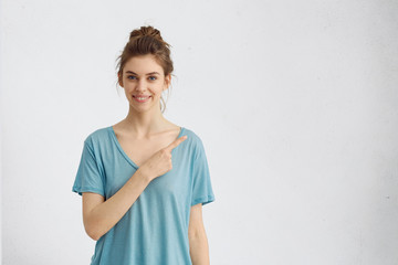 Wall Mural - Isolated studio shot of cheerful young female customer or student in casual blue t-shirt smiling broadly, pointing her index finger at white copyspace wall for your text or advertising information