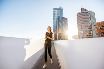 Lifestyle portrait of a stylish woman walking on the modern bridge with skyscrapers on the background during the morning in Amsterdam city