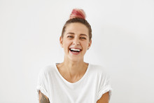 Headshot Of Positive Emotional Gorgeous Young Woman Wearing Pinkish Hair Bun Grinning Broadly, Enjoying Good Day. Beautiful Girl With Tattoo On Arm Having Fun Indoors, Laughing At Something Funny