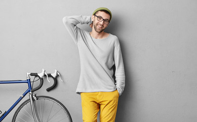 People, travel, leisure and lifestyle concept. Smiling young hipster man in fashionable clothes standing near bike while waiting for his companion to ride together and to explore big city or town