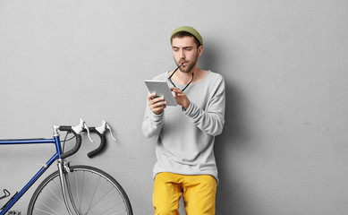 Indoor portrait of handsome male student reading attentively lecture on modern tablet, taking off glasses, being involved in studying, isolated over grey background, standing near his bicycle