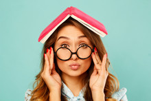 Beautiful Young Student Holding Book On Her Head And Wearing Glasses Over Blue Background