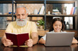 Pleasant grandfather reading book while his granddaughter using laptop