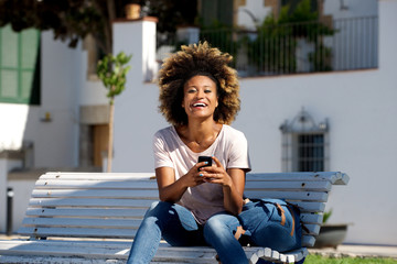 Wall Mural - beautiful young african woman sitting outdoors on bench with mobile phone
