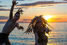 Man With An American Indian Coloring And Feathers On His Head And A Woman Dressed In American Indian Style Performs A Dance On The Seashore At The Sunset