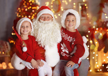 Cute Little Boy And Girl With Santa Claus In Room Decorated For Christmas