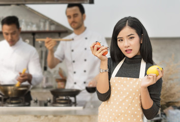 Beautiful woman cooking healthy food in the kitchen.She hold tomatoes and lemon in hands, background is two male chefs are cooking .