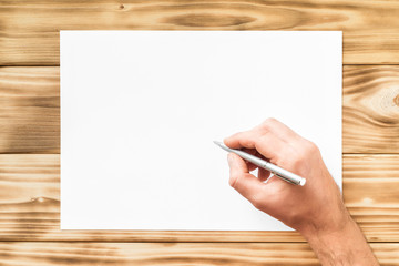 Wall Mural - Mockup of male hand with pen writing at blank white paper list at wooden board background.