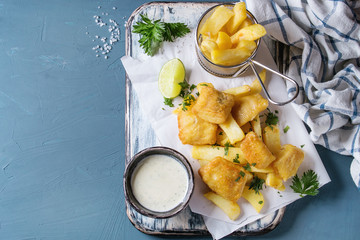 Traditional british fast food fish and chips. Served with white cheese sauce, lime, parsley, french fries in frying basket on white paper over blue concrete background. Top view, copy space