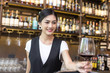 © Bavorndej - Young Asian Woman Waiter Restaurant Catering Service Happy Emotion. Woman Waiter Present Red-Wine for Customer at Bar. Woman with Wine in Bar Concept.