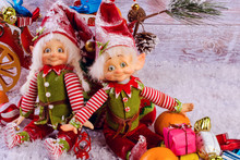 Funny Christmas Elves Sits Next To Christmas Sock On Light Wooden Background. Near Tangerines And Gifts.