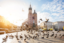 View On The Central Square And Famous St. Marys Basilica With Pigeons Flying During The Sunrise In Krakow, Poland