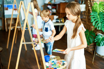 Wall Mural - Group of creative pupils standing at easels with watercolors and paintbrushes in hands and drawing with inspiration, interior of spacious art studio on background