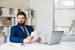 Portrait of successful bearded businessman looking away pensively while working at desk in modern office