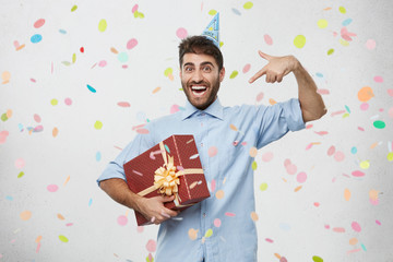 Ecstatic overjoyed young Caucasian man with holiday cap on head holding large box in wrapping paper, feeling excited and euphoric, bragging of his birthday gift, pointing index finger at it