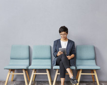 Confident Successful Young Businesswoman With Short Hairstyle Wearing Stylish Clothes And Glasses Using Digital Tablet While Waiting For Doctor's Appointment, Sitting On Chair In Hall At Clinic