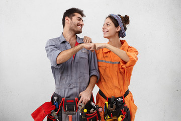 Wall Mural - Teamwork and cooperation concept. Studio shot of young European male and female technicians in overalls standing at blnak wall and making fist bump gesture celebrating good results and success at work