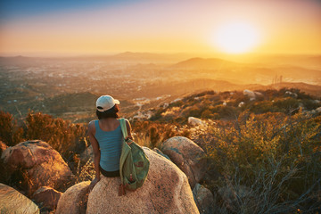 Rear view of woman hiker sitting on rock on top of hill while looking at sunset over San Diego California